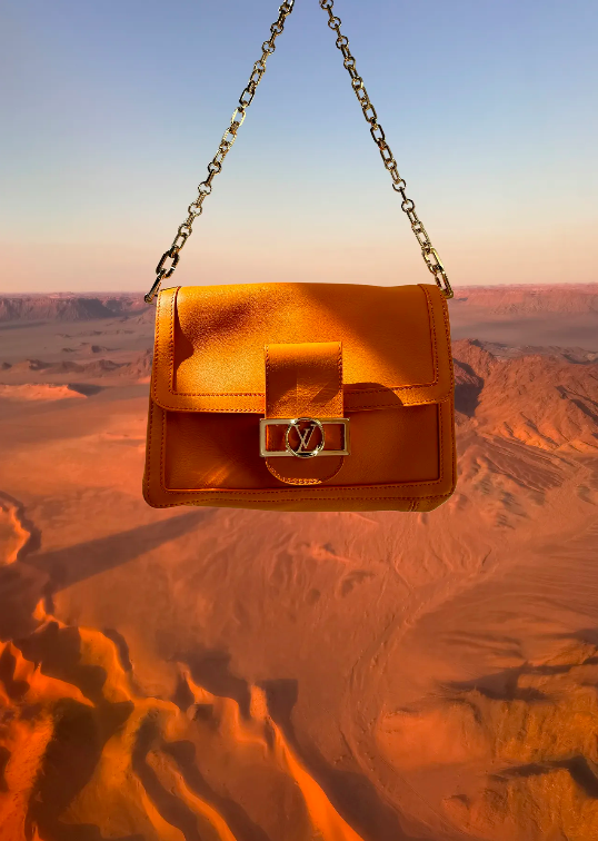 An orange leather purse with gold hardware and chain hangs against a backdrop of sky and sand dunes for a high-fashion product shoot.