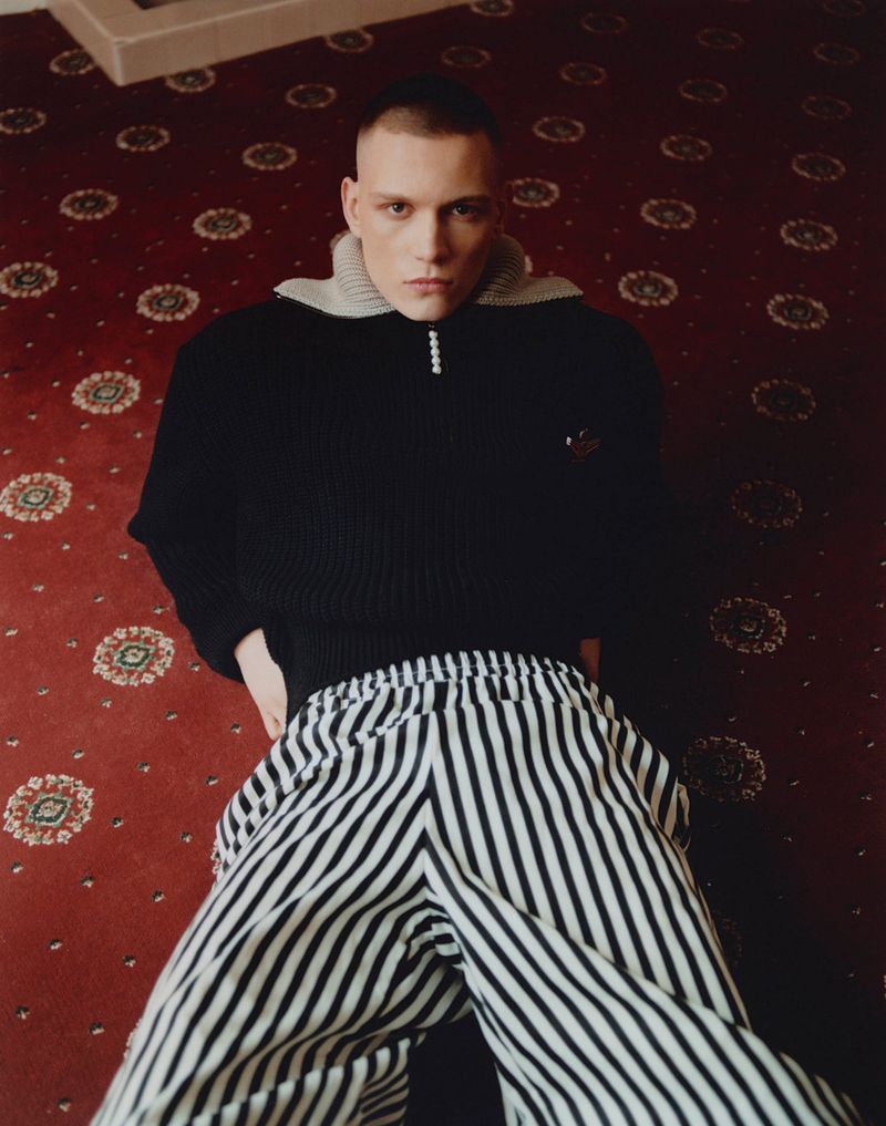 Fashionable adult male poses in a dark sweater and striped pants on a patterned red carpet, looking directly at the camera.