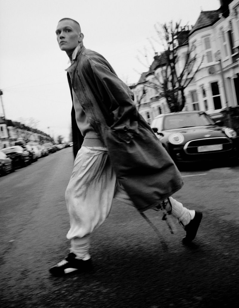 Model Will Woodward walks down a residential street wearing a long coat and loose trousers for The Greatest Magazine editorial.