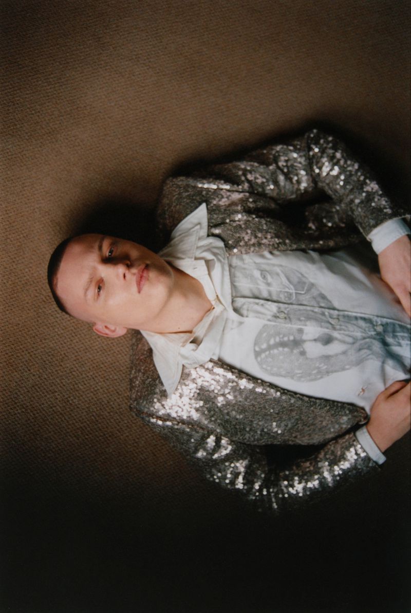 Adult male lying down wearing a sequin jacket and collared shirt with a print pattern on a tan coloured textured surface.