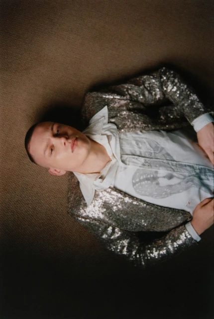 Adult male lying down wearing a sequin jacket and collared shirt with a print pattern on a tan coloured textured surface.