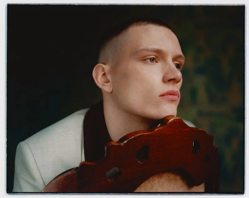 Close-up portrait of a young adult male with a shaved head, wearing a light-colored suit jacket and resting his chin on a wooden chair back.
