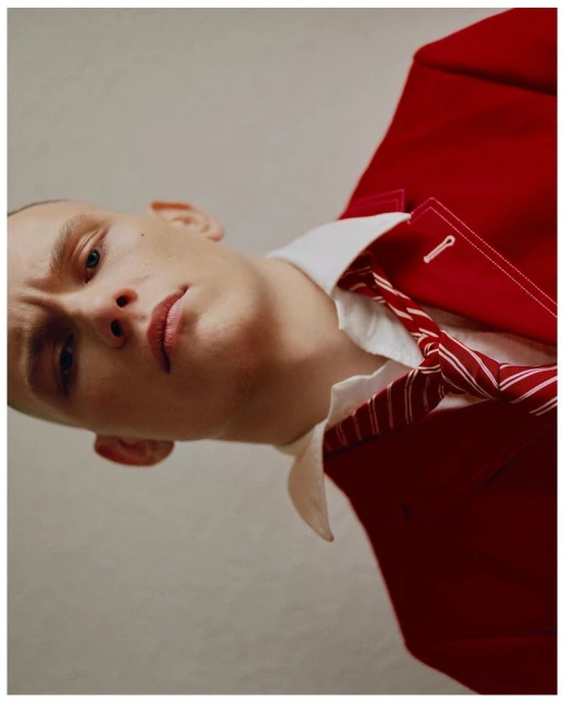 Close-up portrait of a light-skinned adult male with a shaved head, wearing a red suit jacket and a white button-down shirt.