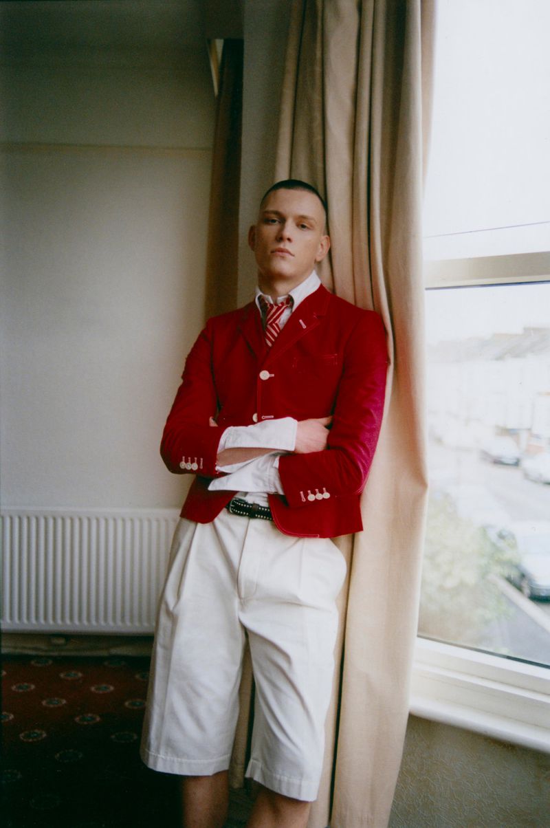 A stylish young adult male with crossed arms poses indoors in a red blazer, white shorts, and a tie, standing near a window with curtains.