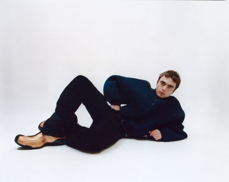 Portrait of a young adult male model laying on a white surface wearing a blue sweater, black pants, and tan shoes.