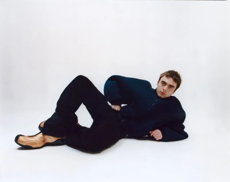 Portrait of a young adult male model laying on a white surface wearing a blue sweater, black pants, and tan shoes.