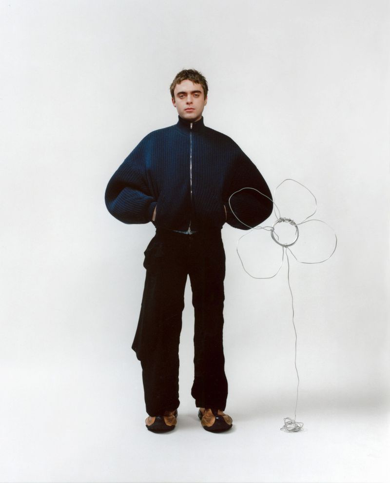 Adult male posing in a navy bomber jacket and black pants, holding a wire flower in a bright photo studio setting.