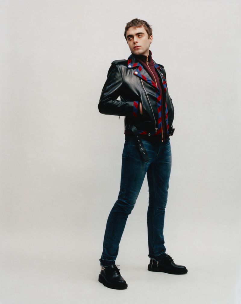 Full-length studio shot of a young adult man wearing a black leather jacket, jeans, and black leather shoes against a plain backdrop.