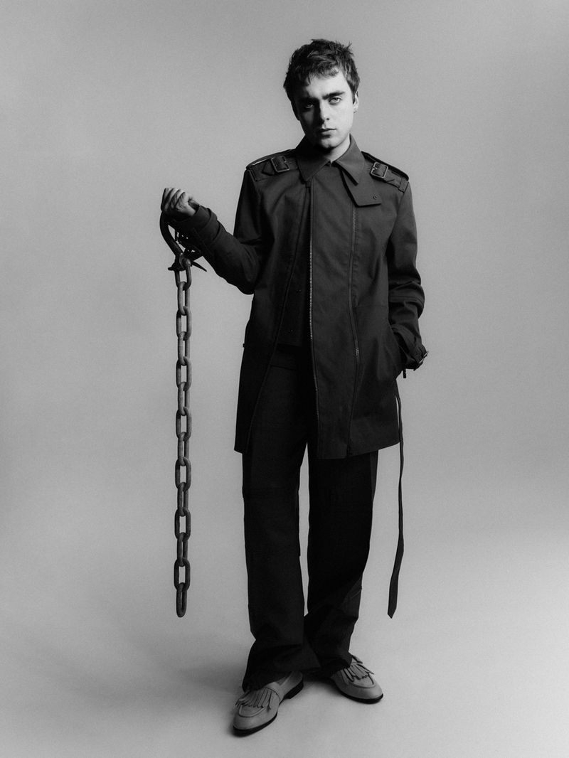 Black and white portrait of an adult male wearing a trench coat and holding a metal chain, standing indoors against a grey backdrop.