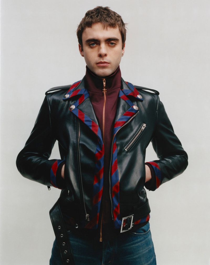 Man with dark hair in leather jacket, maroon turtle neck, and blue jeans posing against a white wall in a studio fashion shot.