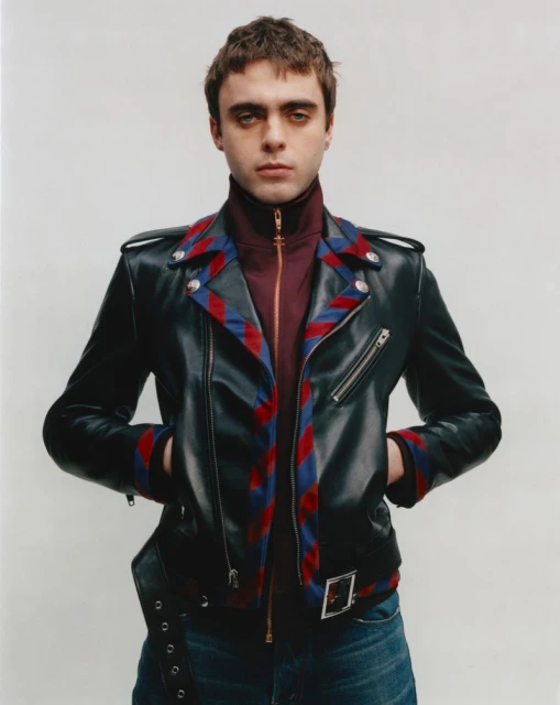 Man with dark hair in leather jacket, maroon turtle neck, and blue jeans posing against a white wall in a studio fashion shot.