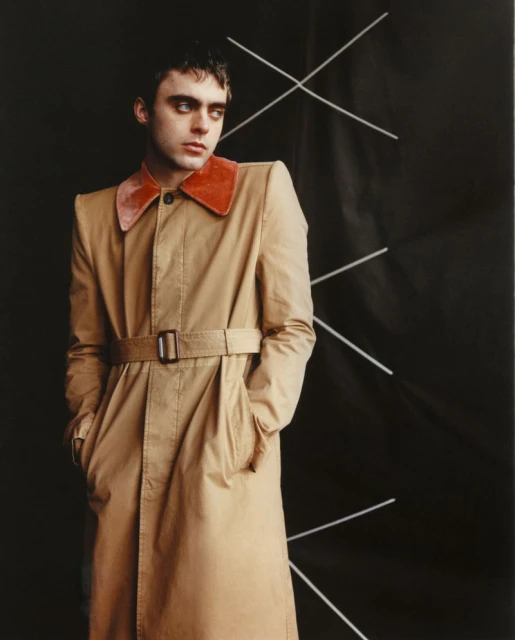 Stylish portrait of a young adult male in a tan trench coat with a red collar, set against a stark black backdrop with white geometric lines.