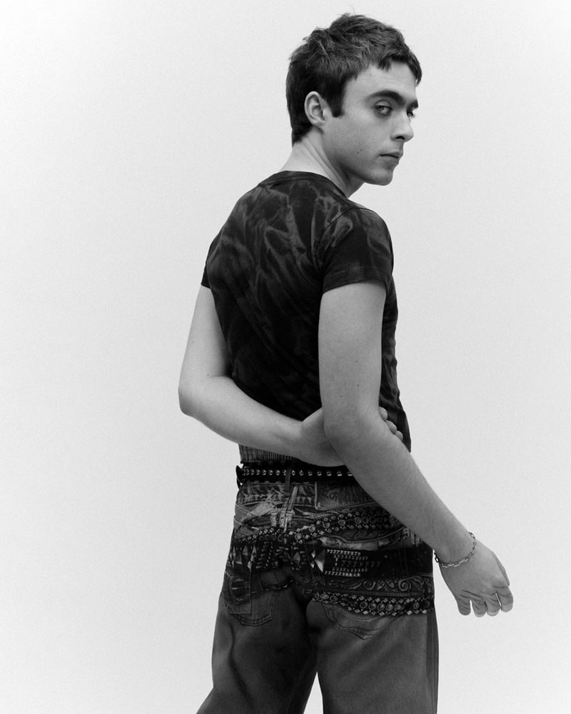 Portrait of a young adult man looking over his shoulder, wearing a black tie-dye t-shirt and decorated jeans, against a plain white background.