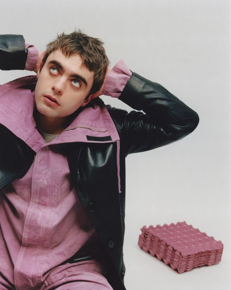 Portrait of a young adult man in a black and lilac jacket looking up with hands behind his head and a stack of egg cartons.