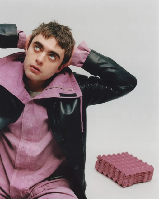 Portrait of a young adult man in a black and lilac jacket looking up with hands behind his head and a stack of egg cartons.