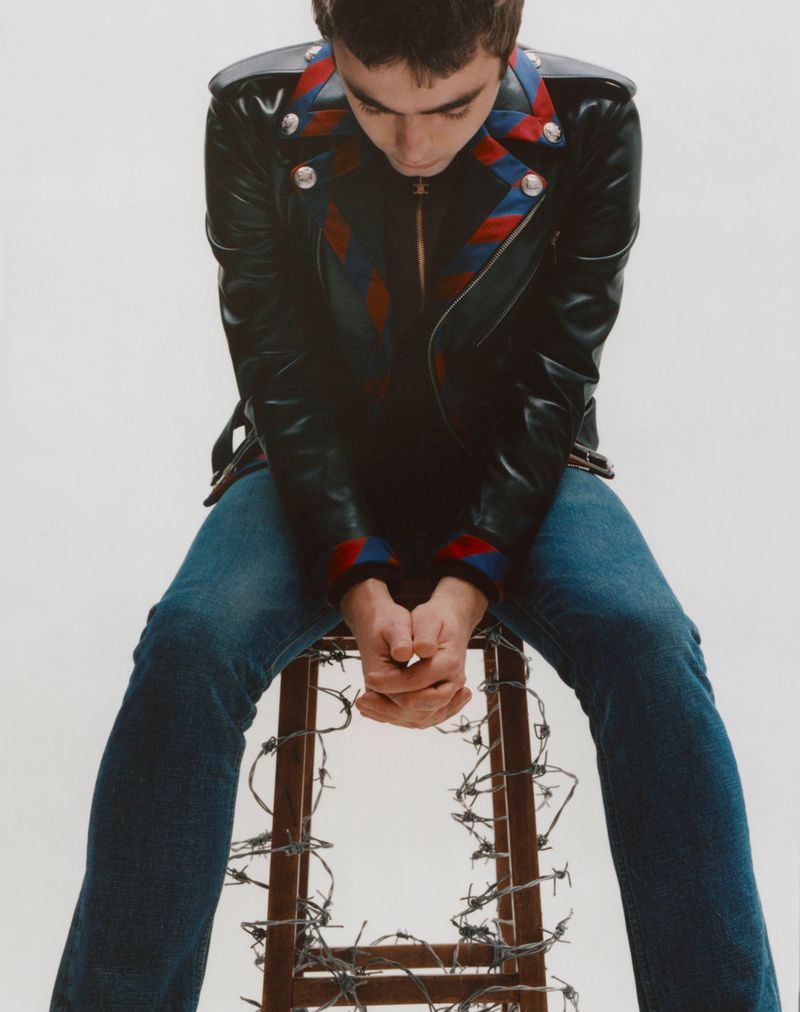 Man in a black leather jacket and jeans sits on a barbed wire-covered stool. Looking down with his hands clasped in an indoor setting.