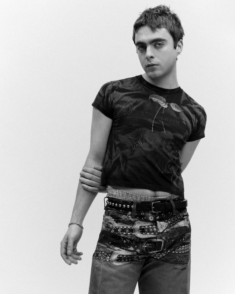 Monochrome portrait of a young man in patterned t-shirt and studded belt posing against a plain white background in studio lighting.