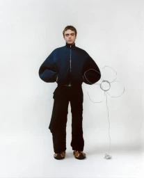 Lennon Gallagher wearing a dark blue zip jacket and black trousers against a plain white seamless background with a wire flower prop.