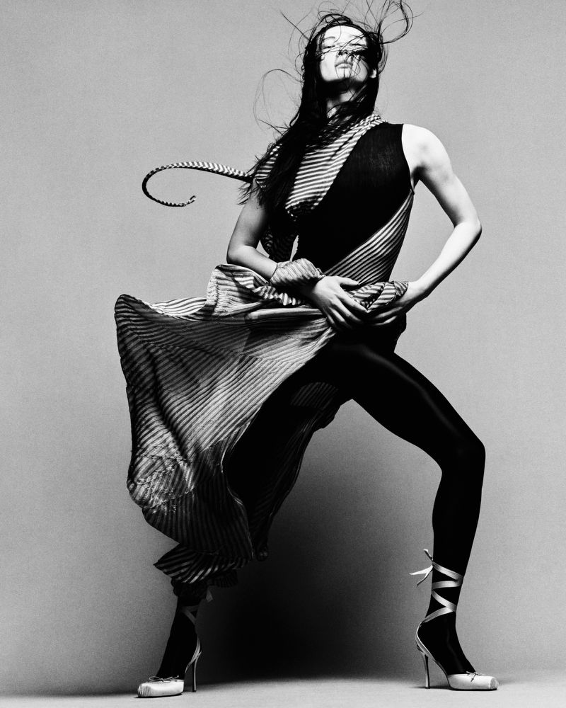 Monochrome full shot of a female fashion model in a flowing striped dress and high heels with ribbon ties, posing on a plain background.
