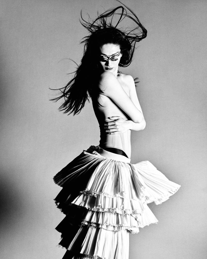 Model with long dark hair blowing upward wearing a structured white tiered skirt against a grey background.