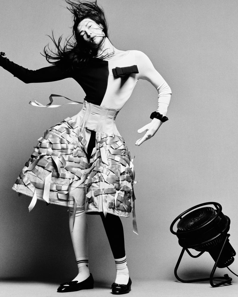 Dramatic black and white fashion shot of a posing woman in an avant-garde ribboned skirt, contrasting top, and theatrical pose.