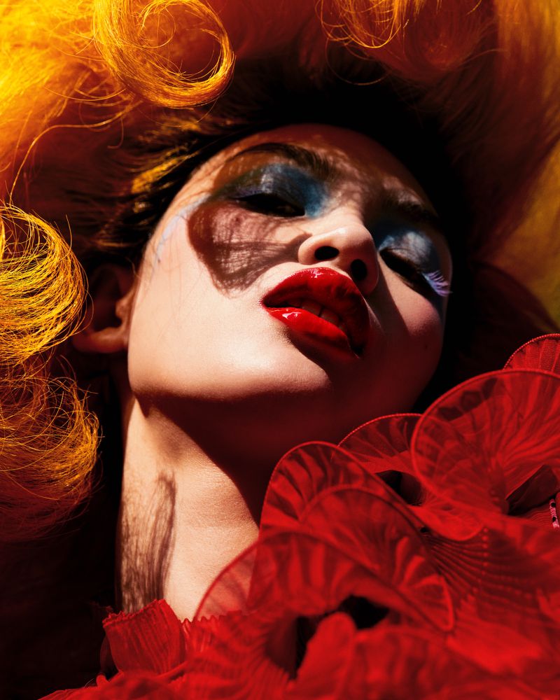 Close-up of an adult woman with vibrant orange hair and dramatic makeup, showcasing red ruffled clothing against a shadowed backdrop.