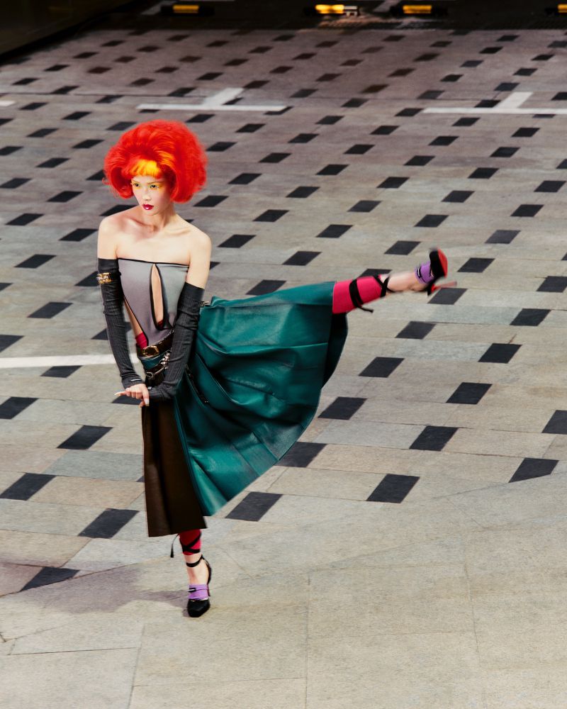 Fashion model with striking red hair strikes a dynamic pose wearing a deconstructed teal dress and black heels on a patterned floor.