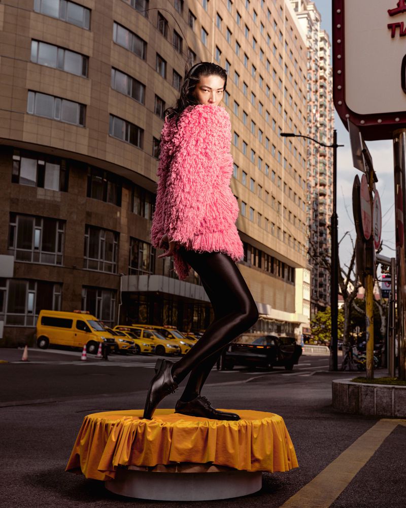 A woman in a pink faux fur coat and shiny black leggings poses on a yellow pedestal in an urban setting with tall buildings and taxis.