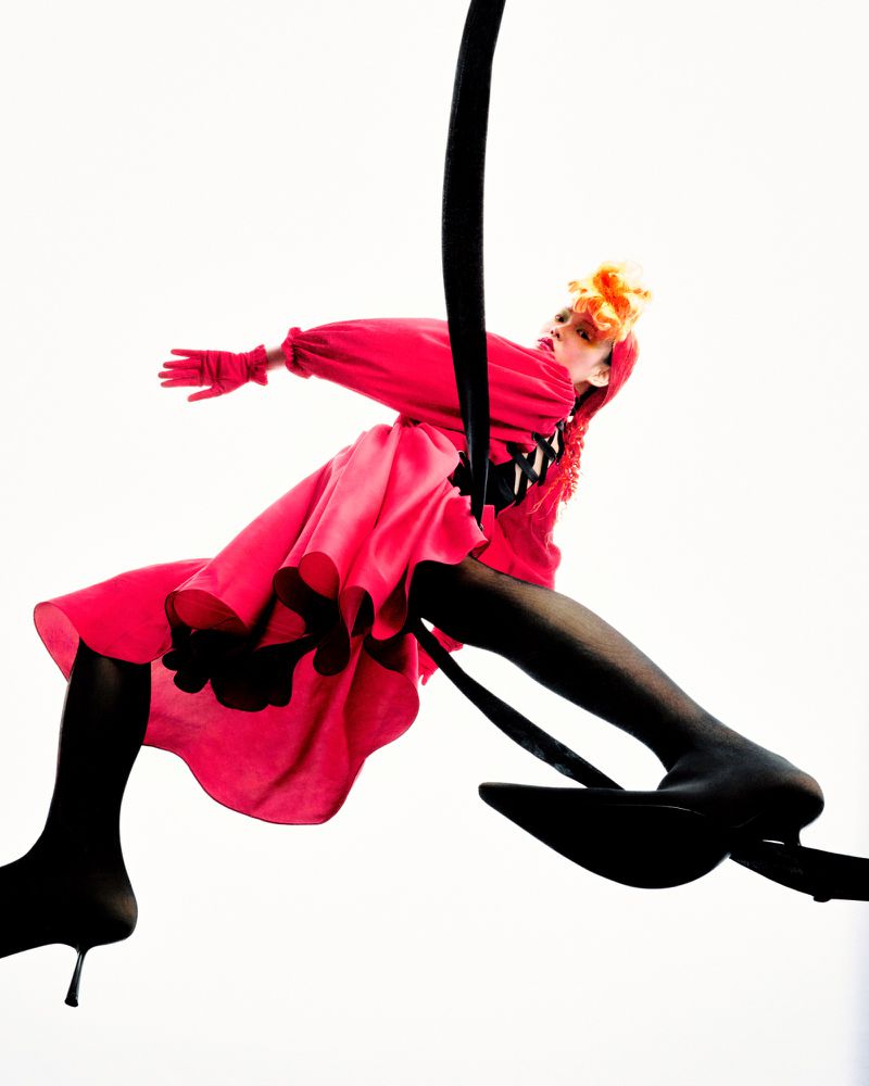 A woman in a pink dress and black tights is suspended from a black strap against a white background in a dramatic fashion shot.