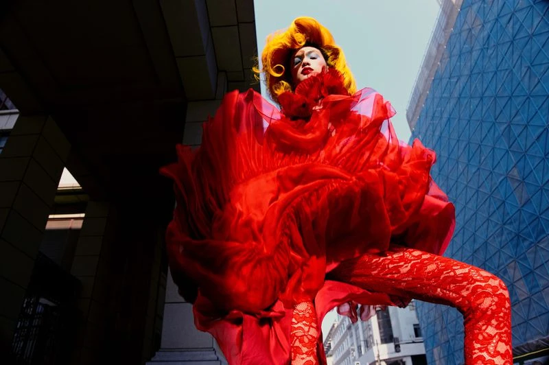 Dramatic fashion portrait of a person in an oversized red dress with yellow hair standing in a city street, shot from a low angle.