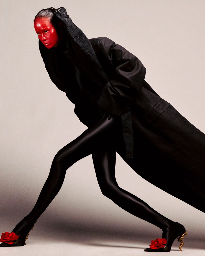 Person with face painted red and dressed in black coat, leggings and high heels shoes with decorative red rose posing indoors.
