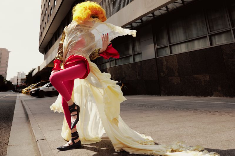 Fashionable person in bright colors poses on city street near buildings and parked cars with flowing fabric accentuating their look.
