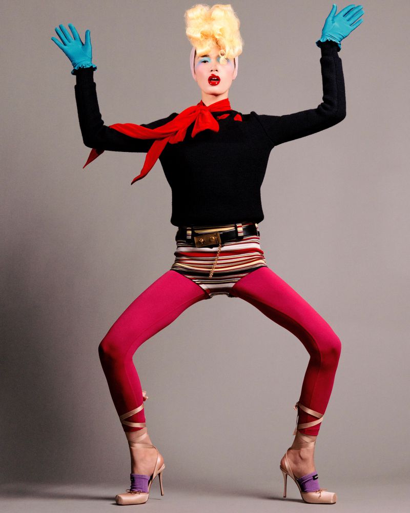A female fashion model poses in a black sweater, pink leggings, and high heels against a gray background in a studio setting.
