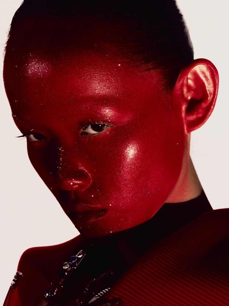 Close-up portrait of a woman with red painted skin and glitter details, wearing a red top against a light background. Beauty shot.