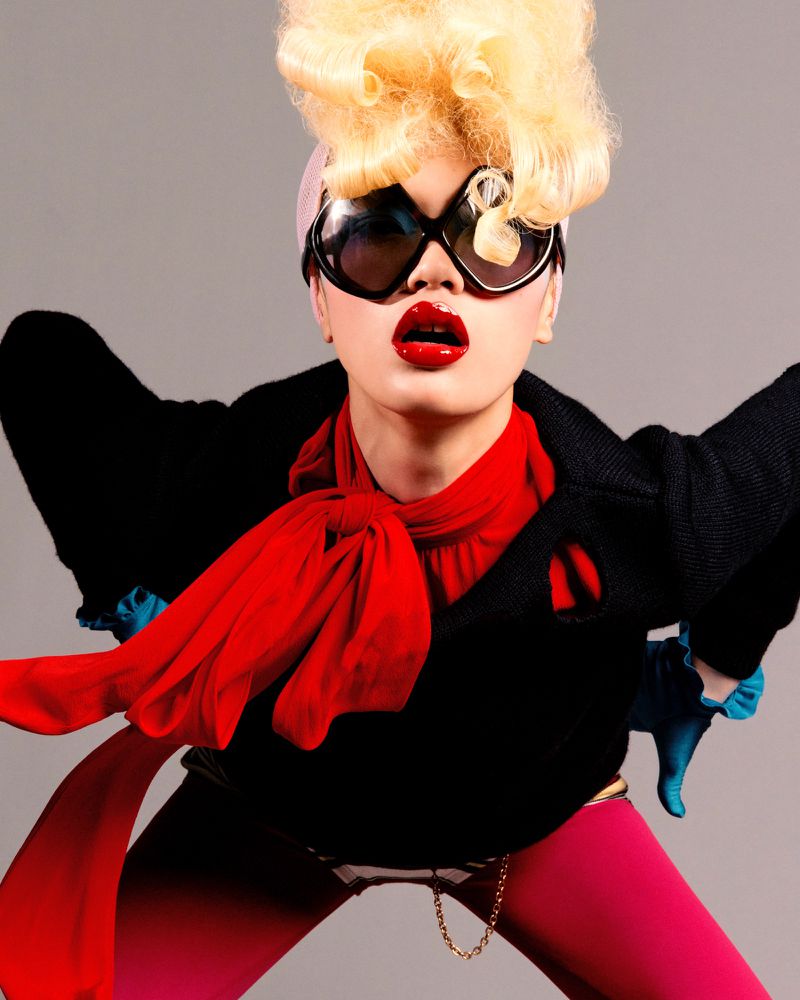 Fashion photo of a woman with a bouffant hairstyle, dark sunglasses, red lips, black blazer, and bright red scarf posing in a studio.