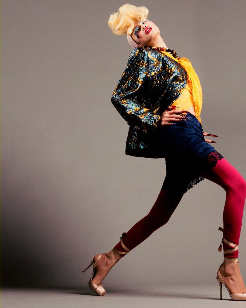 Full-length fashion shot of a woman with a blonde wig, pink and red tights, and heels, posing against a gray background in a studio.
