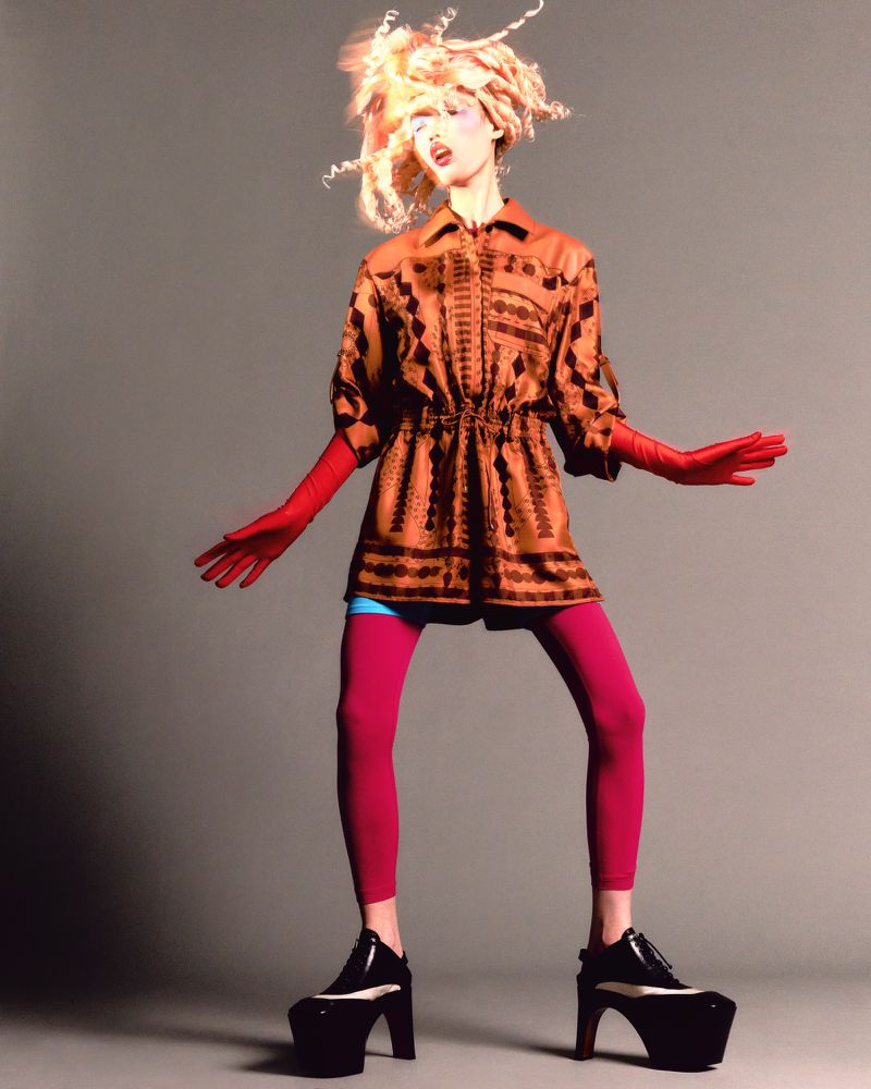 Fashion model strikes a pose in a short patterned shirt dress, red leggings, red gloves and large black platform shoes against grey.