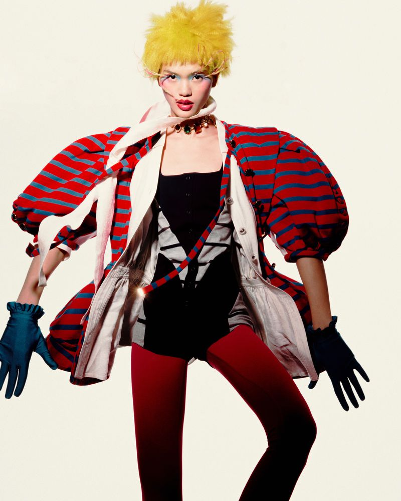 Fashion editorial shot of a woman with vibrant yellow hair wearing a striped jacket, black leotard, red tights, and blue gloves in a studio.