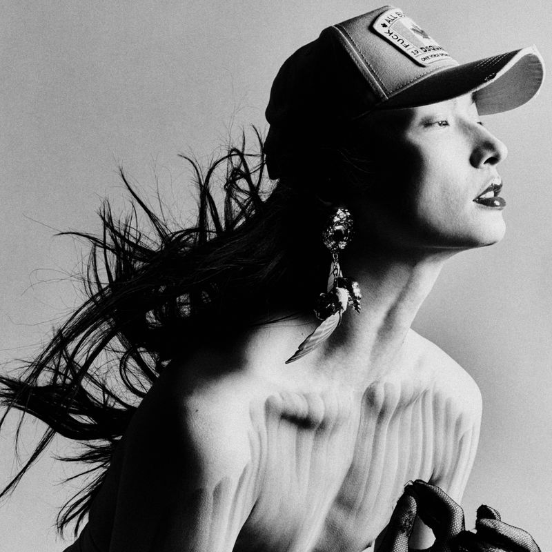 Black and white portrait of a stylish adult woman wearing a baseball cap, large earrings and dramatic makeup looking away from the camera.