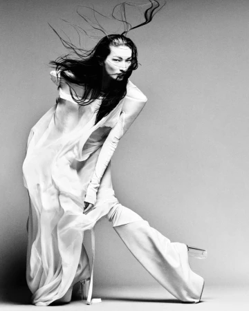 Model in flowing white garments posing dynamically with long dark hair blowing against a grey background