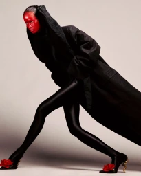 Model with painted red face wearing a black coat and tights with red floral pumps against a grey seamless background