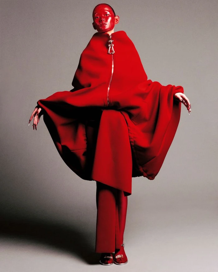 Model with bright red painted face wearing a sculptural red cape and trousers against a grey backdrop