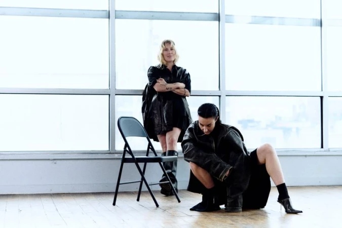 Two women pose in an indoor studio setting with large windows, dressed in dark, fashionable clothing next to a black folding chair.