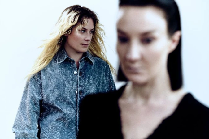 Two women pose in front of a white background. One woman with blonde hair is in focus; the other woman is blurred in the background.