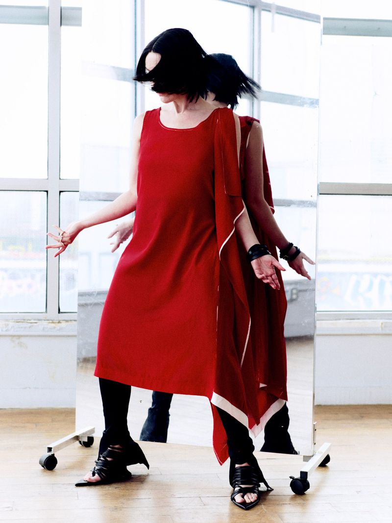 A woman with short black hair poses in a red dress and black leggings, reflected in a mirror. Window and light wood floor background.