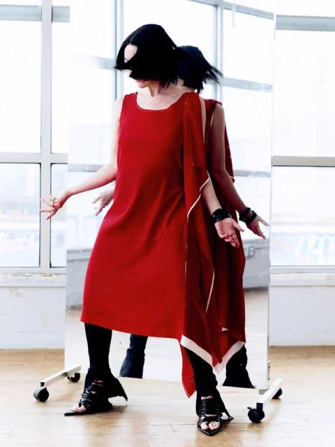 A woman with short black hair poses in a red dress and black leggings, reflected in a mirror. Window and light wood floor background.