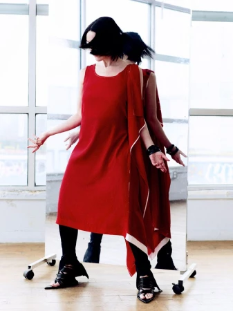 A woman wearing a bright red dress stands in front of a rolling mirror reflecting her back.
