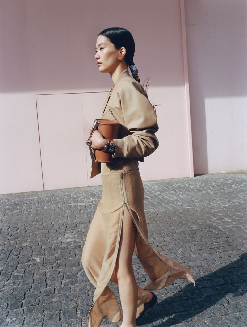 Asian woman in a tan jacket and long beige skirt walking on a cobblestone street with a pink wall while holding a brown handbag.