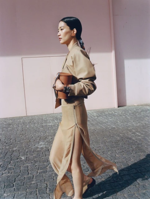 Asian woman in a tan jacket and long beige skirt walking on a cobblestone street with a pink wall while holding a brown handbag.