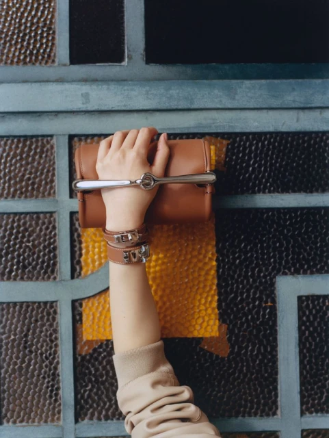 Tan leather purse held by a child's arm with bracelets, angled view against a gridded window backdrop with blue and amber.